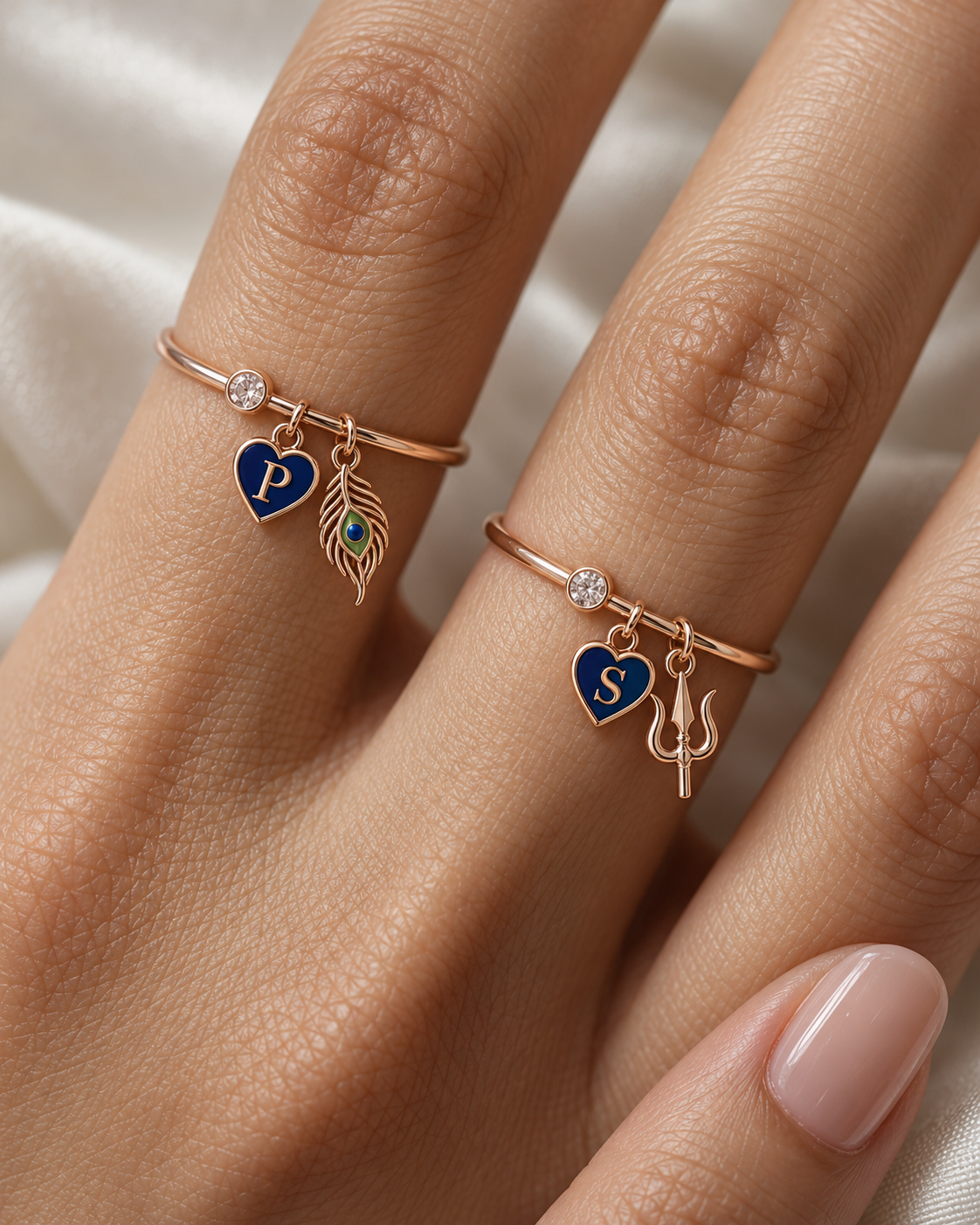 Hari Har Neelam Lab Diamond Silver Couple Rings – pair of rose gold tone charm rings with navy-blue enamel heart initials, a gold peacock-feather charm (Krishna) and a gold trishul charm (Shiva), lab grown diamond solitaires, worn on the hand