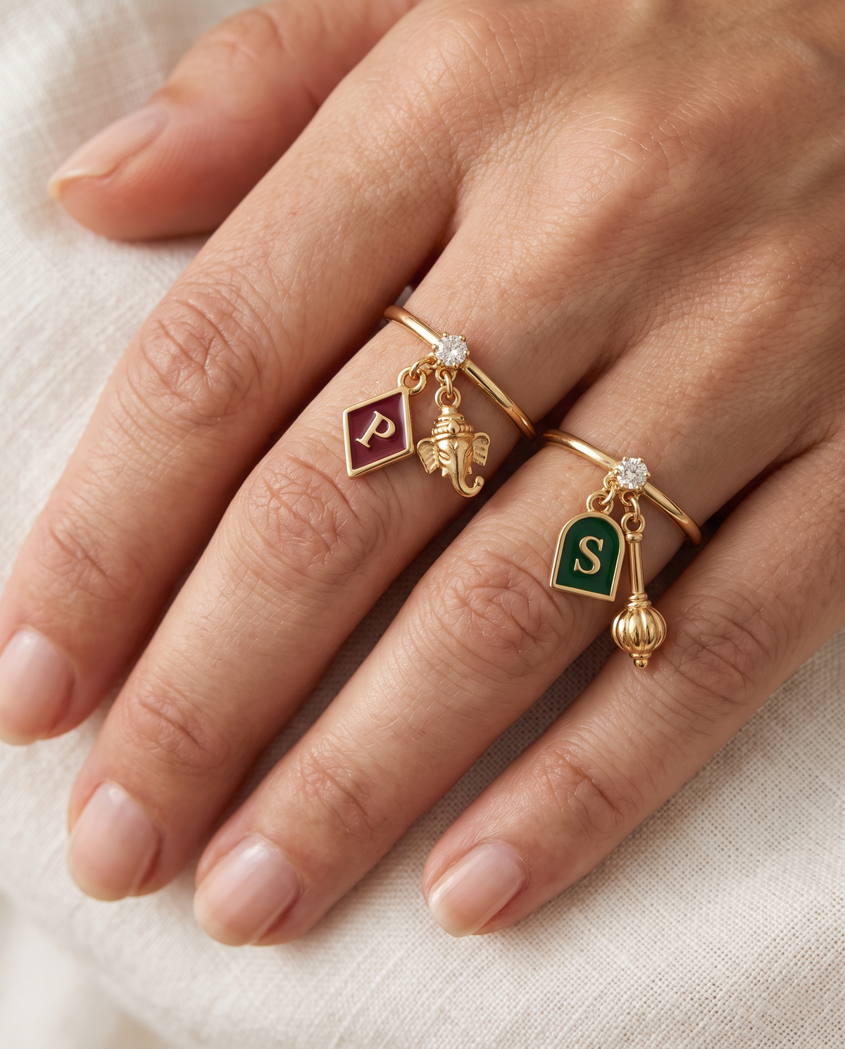 Gajanan Pawanputra Lab Diamond Silver Couple Rings – pair of gold tone charm rings with maroon enamel diamond-shaped initial and gold Ganesha charm, and emerald-green enamel arch initial and gold gada charm (Hanuman), lab grown diamond solitaires, worn on the hand